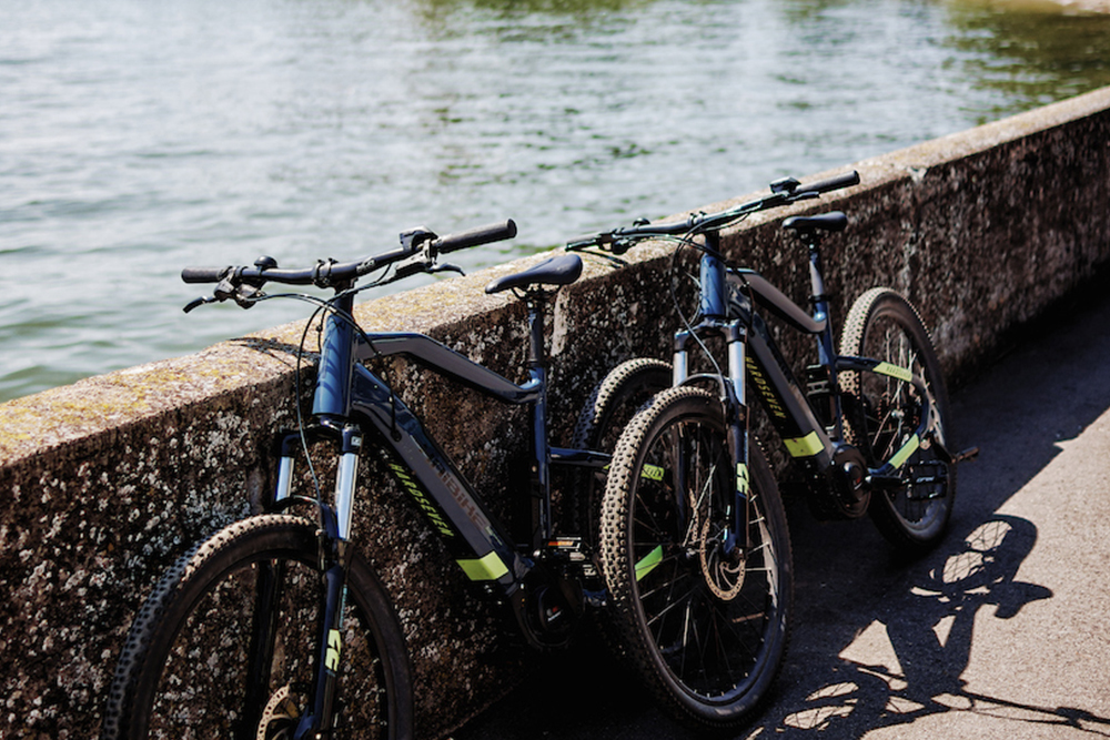 Two e-bikes lean against a bank wall with a view of Lake Constance in the sunshine.