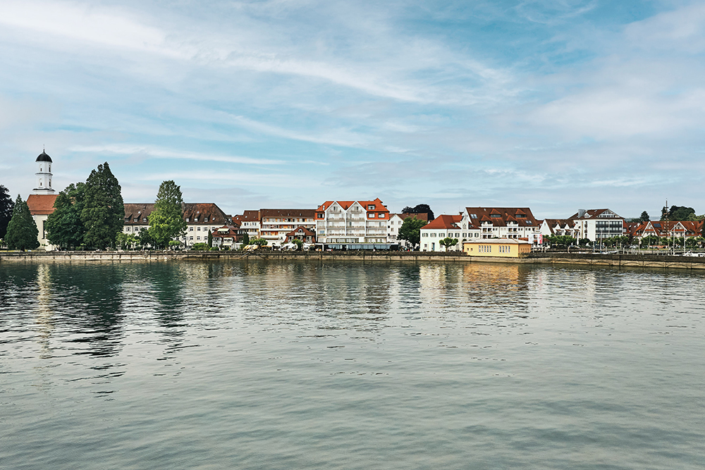 Panoramic view of a lakeside town on Lake Constance with a church, hotels and reflecting water.