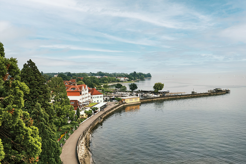 Aerial view of the Lake Constance promenade with buildings, harbor facilities and a wide view over the lake.