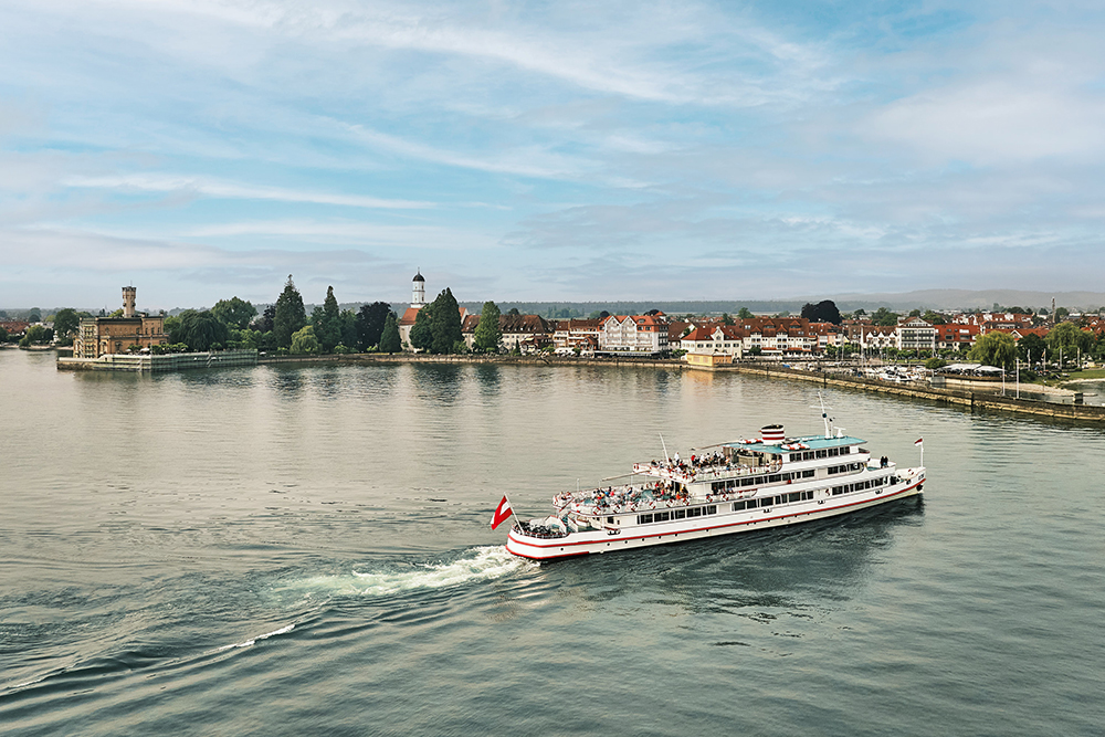 A passenger ship sails across Lake Constance in front of a lakeside town with historic buildings and calm waters.