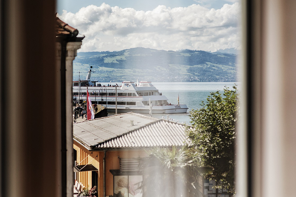 View through a window of a passenger ship in the harbor of Lake Constance against a mountainous backdrop.
