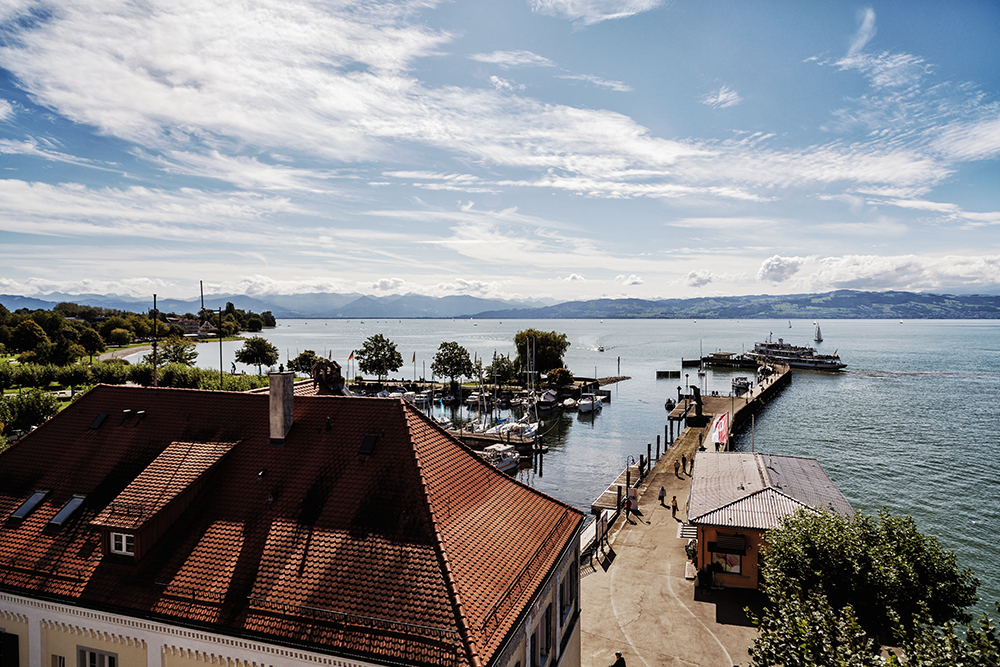 View over the harbor on Lake Constance with boats, jetty and mountains in the background under a blue sky.