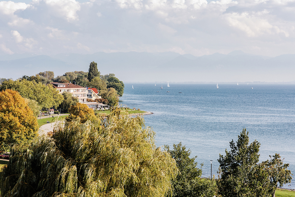 Panoramic shot of the shore of Lake Constance with sailing boats, trees and mountains in the background.
